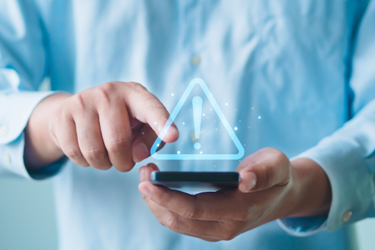 Gros plan sur des mains tenant un téléphone intelligent affichant un symbole d’alerte.