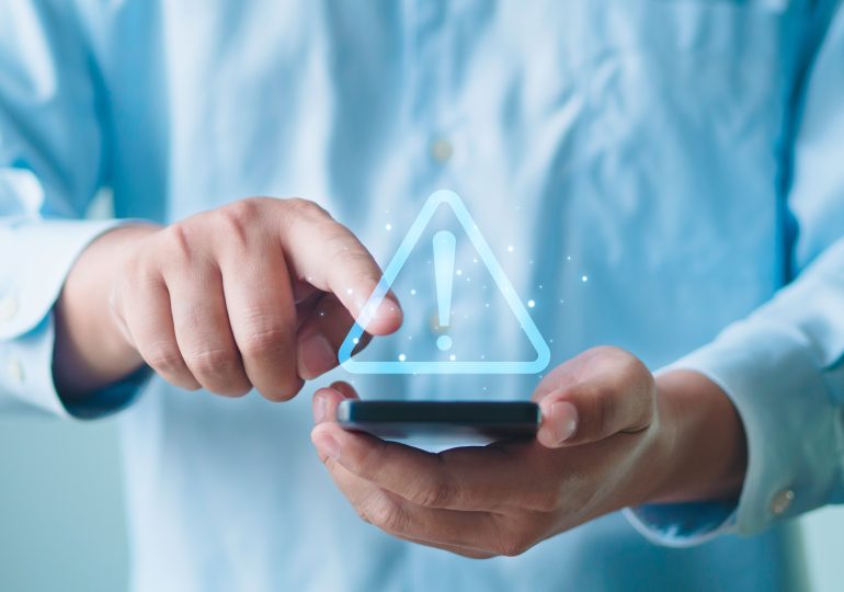 Gros plan sur des mains tenant un téléphone intelligent affichant un symbole d’alerte.
