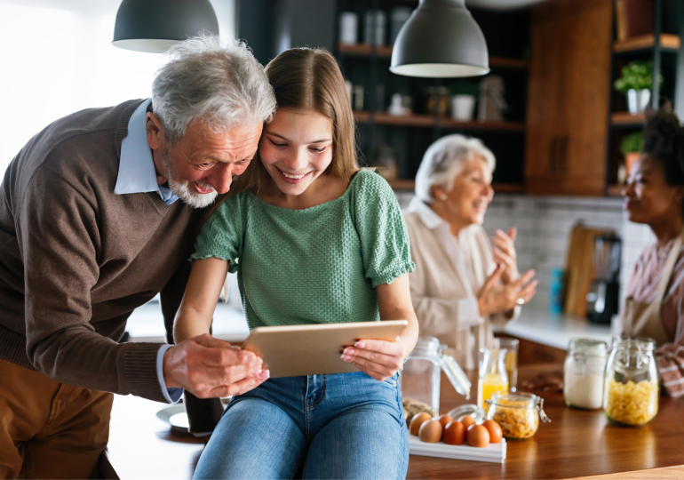 Un homme et une femme consultant une tablette dans une cuisine, tandis que deux femmes discutent en arrière‑plan.