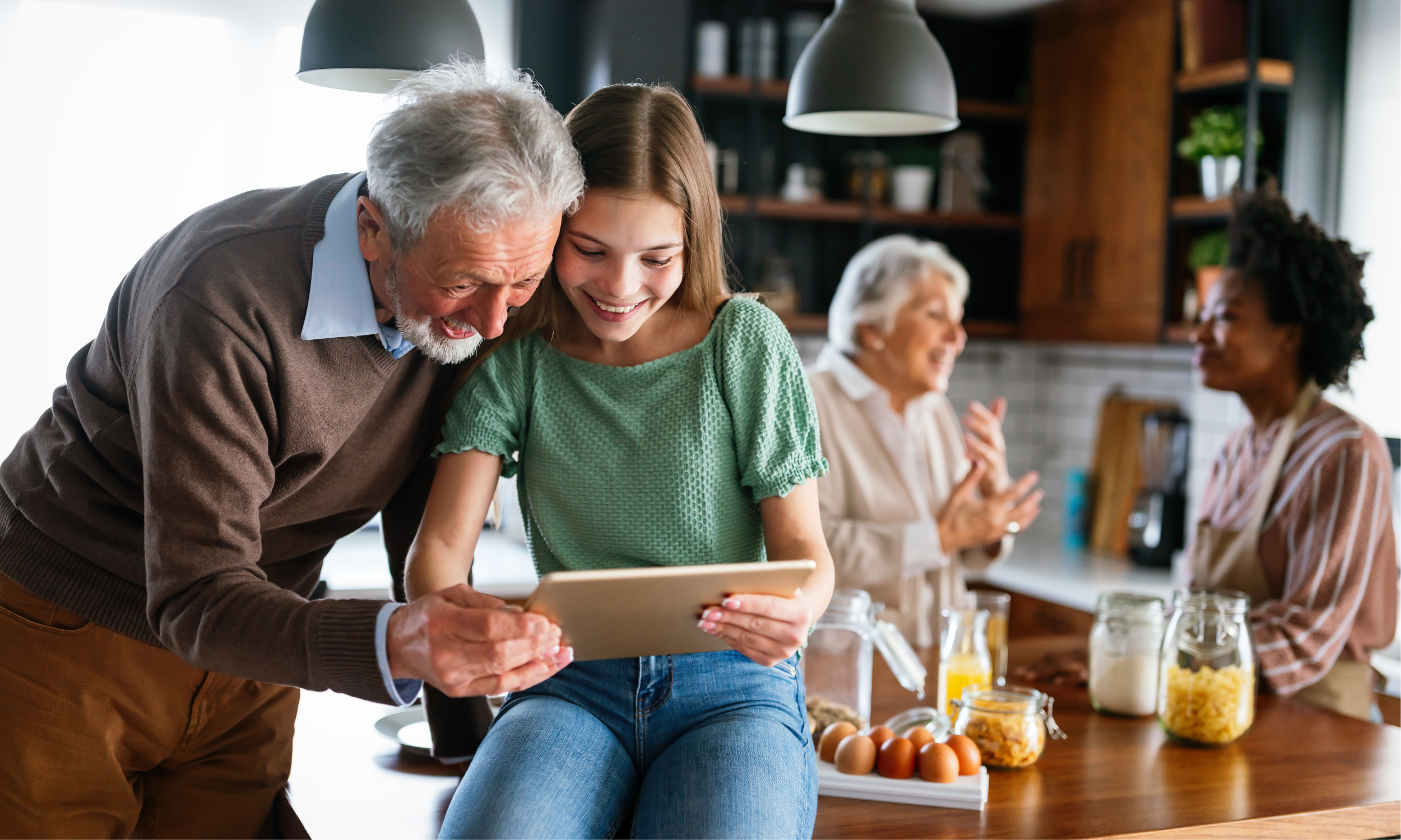 Un homme et une femme consultant une tablette dans une cuisine, tandis que deux femmes discutent en arrière‑plan.